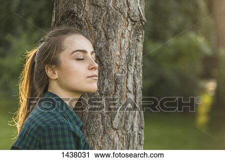 Stock Image - Beautiful woman with eyes closed leaning on tree trunk at park. Fotosearch
