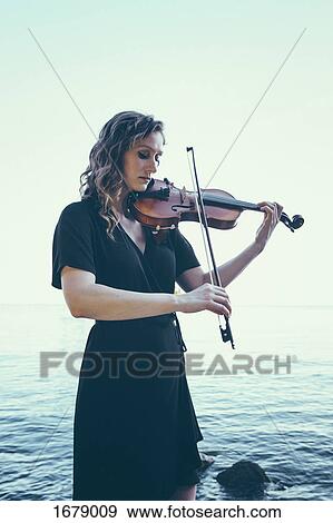 Stock Photo - Beautiful young woman playing violin while standing by lake against clear sky. Fotosearch