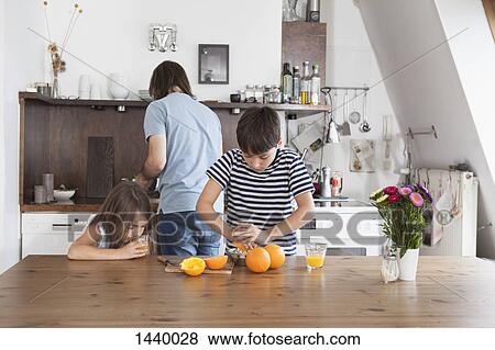 Stock Photo - Boy squeezing oranges while sister drinking juice with father in background at kitchen. Fotosearch