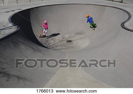 Stock Image - Brother and sister on scooters playing at sunny skate park. Fotosearch