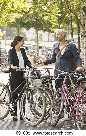 Stock Photography - Business colleagues talking while standing with bicycles in parking lot. Fotosearch