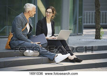 Stock Photograph - Business coworkers with laptop discussing while sitting on steps in city. Fotosearch