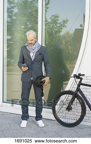Stock Photography - Businessman using mobile phone while standing by bicycle against glass wall. Fotosearch