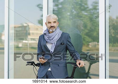 Picture - Businessman with bicycle holding smart phone while standing against glass wall. Fotosearch