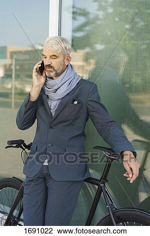 Businessman with bicycle talking on mobile phone while standing against glass building View Large Photo Image Stock Image - Businessman with bicycle talking on mobile phone while standing against glass building. Fotosearch
