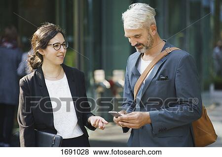 Businesswoman looking at male colleague using mobile phone while standing in city View Large Photo Image Stock Photo - Businesswoman looking at male colleague using mobile phone while standing in city. Fotosearch