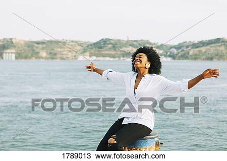 Stock Image - Carefree young woman with arms outstretched at waterfront, Belem, Lisbon, Portugal. Fotosearch