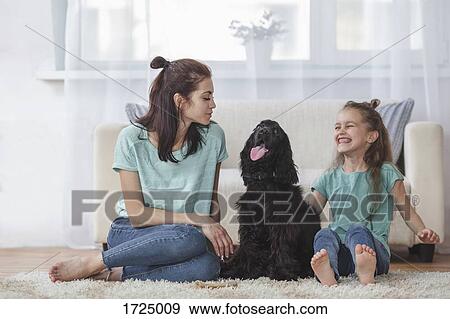 Stock Photo - Cheerful girl sitting by Cocker Spaniel and mother in living room at home. Fotosearch