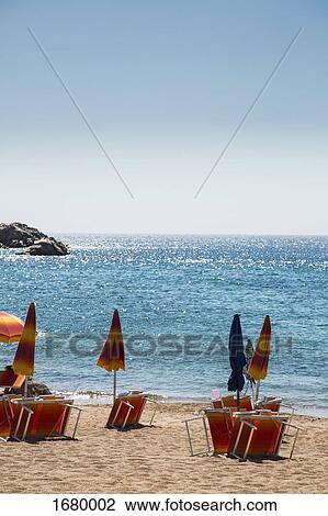 Stock Image - Closed parasols with deck chairs at beach against clear blue sky on sunny day. Fotosearch