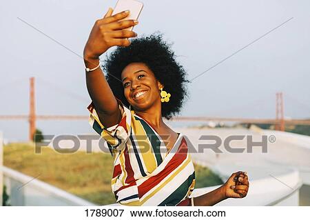 Confident young woman taking selfie with smart phone with 25 de Abril Bridge in background View Large Photo Image Stock Photo - Confident young woman taking selfie with smart phone with 25 de Abril Bridge in background. Fotosearch