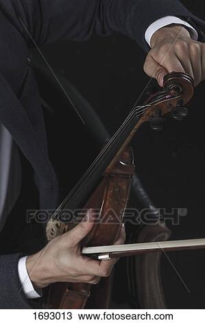 Stock Image - Cropped image of violinist tuning violin while sitting on chair. Fotosearch