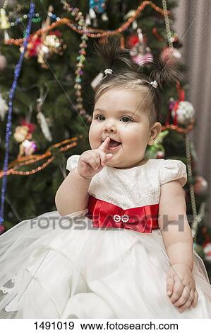 Stock Photo - Cute baby girl sitting with Christmas tree in background. Fotosearch