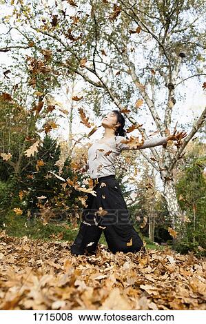 Dry leaves falling over young woman standing on field against trees at park View Large Photo Image Stock Photo - Dry leaves falling over young woman standing on field against trees at park. Fotosearch