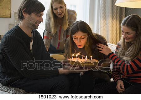Stock Image - Family looking at young woman blowing out birthday candles at home. Fotosearch
