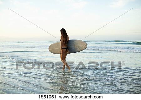 Stock Image - Female surfer carrying surfboard in ocean surf. Fotosearch