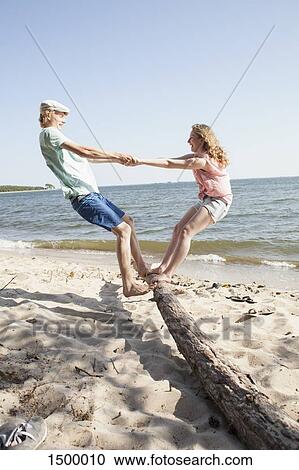 Stock Image - Friends enjoying while standing on driftwood at beach against clear sky. Fotosearch