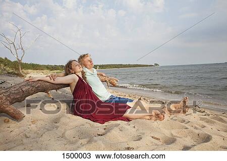 Stock Image - Friends relaxing while leaning on driftwood at beach against sky. Fotosearch