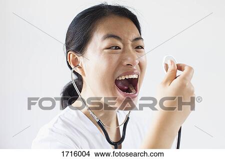 Picture - Frustrated doctor screaming with mouth open into stethoscope against white wall. Fotosearch