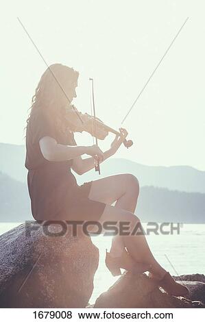 Full length of backlit woman playing violin while sitting on rock at lakeshore against clear sky View Large Photo Image Stock Photo - Full length of backlit woman playing violin while sitting on rock at lakeshore against clear sky. Fotosearch