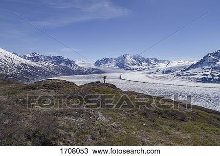 Stock Image - Full length of hiker looking at glacier against blue sky, Knik Glacier, Palmer, Alaska. Fotosearch