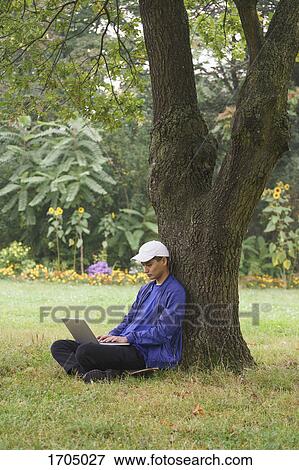 Full length of man using laptop while sitting by tree on grassy field at park View Large Photo Image Stock Photo - Full length of man using laptop while sitting by tree on grassy field at park. Fotosearch