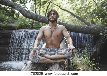 Stock Photography - Full length of shirtless man sitting with crossed legs on logs against waterfall at forest. Fotosearch