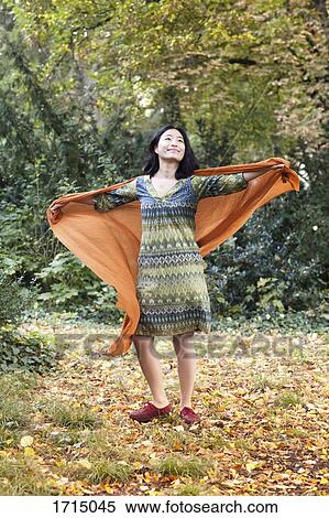 Stock Photography - Full length of woman wearing orange scarf standing with arms outstretched at park during autumn. Fotosearch