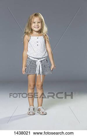 Stock Image - Full length portrait of happy girl standing against gray wall. Fotosearch