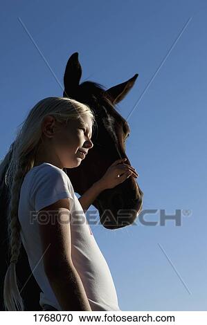 Stock Image - Girl petting horse muzzle under blue sky. Fotosearch