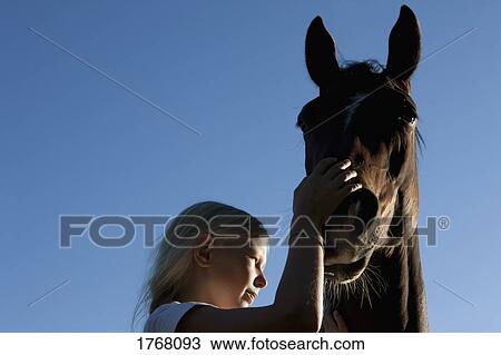 Stock Image - Girl petting muzzle of horse under blue sky. Fotosearch