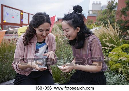 Stock Photograph - Happy female friends using smart phones while sitting on patio. Fotosearch