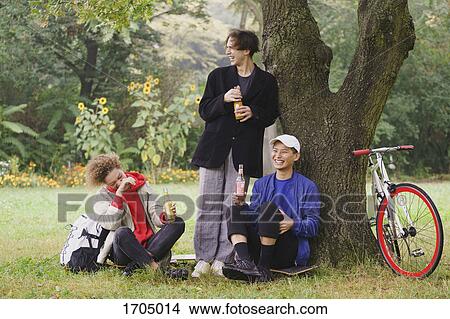 Happy friends drinking juice while relaxing below tree on grassy field at park View Large Photo Image Picture - Happy friends drinking juice while relaxing below tree on grassy field at park. Fotosearch