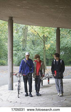 Stock Image - Happy friends talking while walking in gazebo against trees at park. Fotosearch