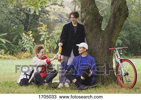 Happy friends toasting drinks while relaxing below tree on grassy field at park View Large Photo Image Stock Image - Happy friends toasting drinks while relaxing below tree on grassy field at park. Fotosearch