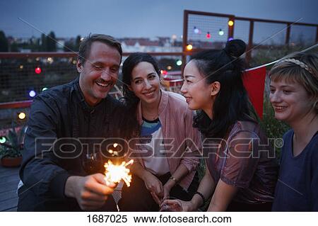 Stock Photography - Happy man holding sparkler while sitting with female friends at night. Fotosearch