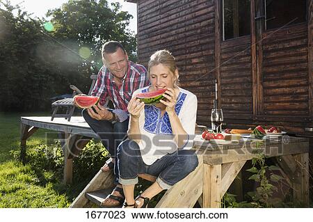 Stock Image - Happy mature couple enjoying watermelon slice while sitting outside farmhouse on sunny day. Fotosearch