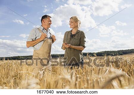 Stock Image - Happy mature couple talking while standing amidst crops at farm against sky. Fotosearch