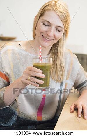 Happy mature woman holding a smoothie in a glass at home View Large Photo Image Picture - Happy mature woman holding a smoothie in a glass at home. Fotosearch