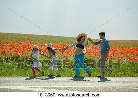 Stock Image - Happy pregnant family holding hands, walking along sunny rural red poppy field. Fotosearch