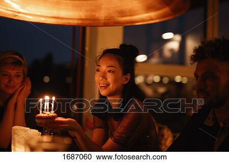 Happy woman holding small birthday cake with lit candles while sitting by friends at home View Large Photo Image Stock Image - Happy woman holding small birthday cake with lit candles while sitting by friends at home. Fotosearch