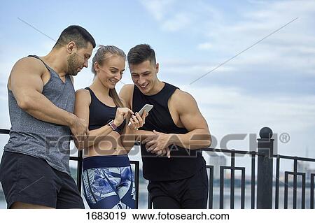 Stock Photo - Happy young athletes using mobile phone while leaning by railing. Fotosearch