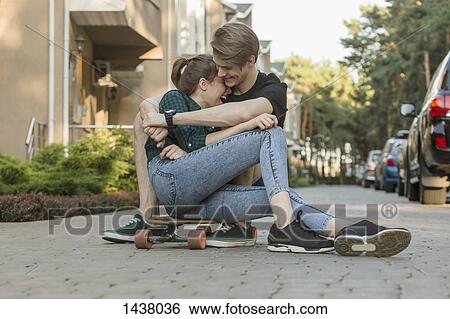Stock Photograph - Happy young couple embracing while sitting on skateboard outdoors. Fotosearch