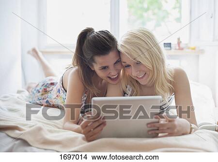 Picture - Happy young female friends using digital tablet while lying on bed at home. Fotosearch