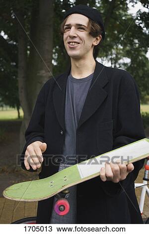 Stock Image - Happy young man holding skateboard while standing at park. Fotosearch
