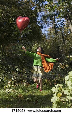 Happy young woman holding red heart shape balloon while running against trees at park View Large Photo Image Stock Image - Happy young woman holding red heart shape balloon while running against trees at park. Fotosearch