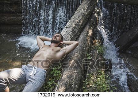 Stock Photo - High angle view of thoughtful shirtless man lying on logs over waterfall at forest. Fotosearch