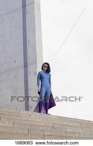 Stock Image - Low angle view of confident female superhero standing on steps against clear sky. Fotosearch