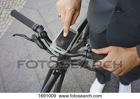 Stock Photo - Low section of businessman using smart phone on bicycle handle while standing at street. Fotosearch