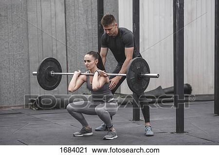 Stock Image - Male instructor assisting young woman crossfit training at gym. Fotosearch