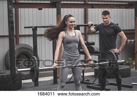 Stock Image - Male instructor looking at female athlete smiling while lifting barbell during cross training. Fotosearch